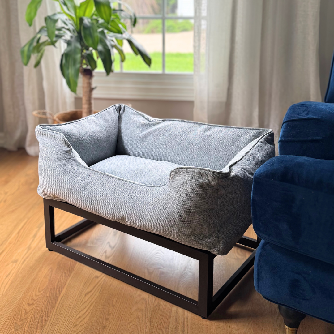 Gray pet bed on a wooden floor with a blue armchair and plant in the background