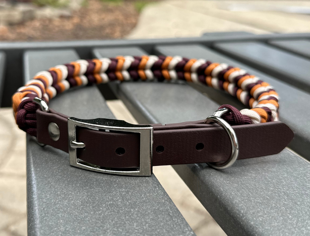 Dog collar with multicolored braided design and brown leather section on a metal surface.