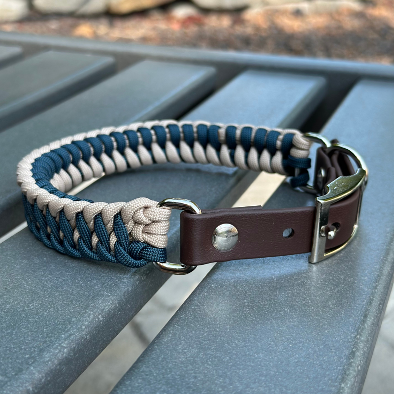 Dog collar with navy blue and light tan braided design with a dark brown BioThane strap belt buckle style silver hardware, sitting on a gray bench