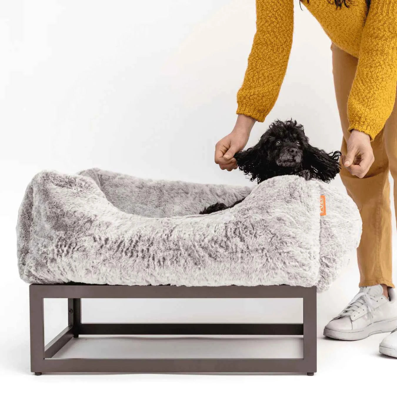 The FÜZI Dog Bed in gray on top of frame with small black dog on white background with woman in yellow outfit petting the dog's ears