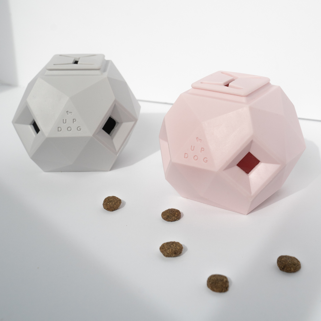 Two geometric dog toys, one white and one pink, on a light surface with scattered dog treats.