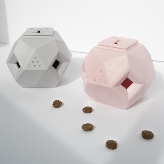 Two geometric dog toys, one white and one pink, on a light surface with scattered dog treats.