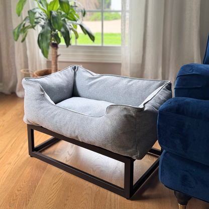 Gray pet bed on a wooden floor with a blue armchair and plant in the background