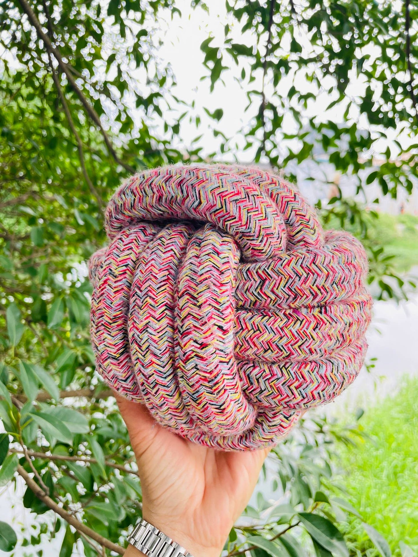 Multicolored rope knot held by a hand with a green leafy background