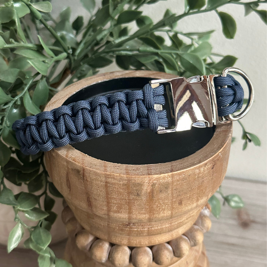 Blue paracord dog collar with silver buckle on a wooden stand with green leaves in the background