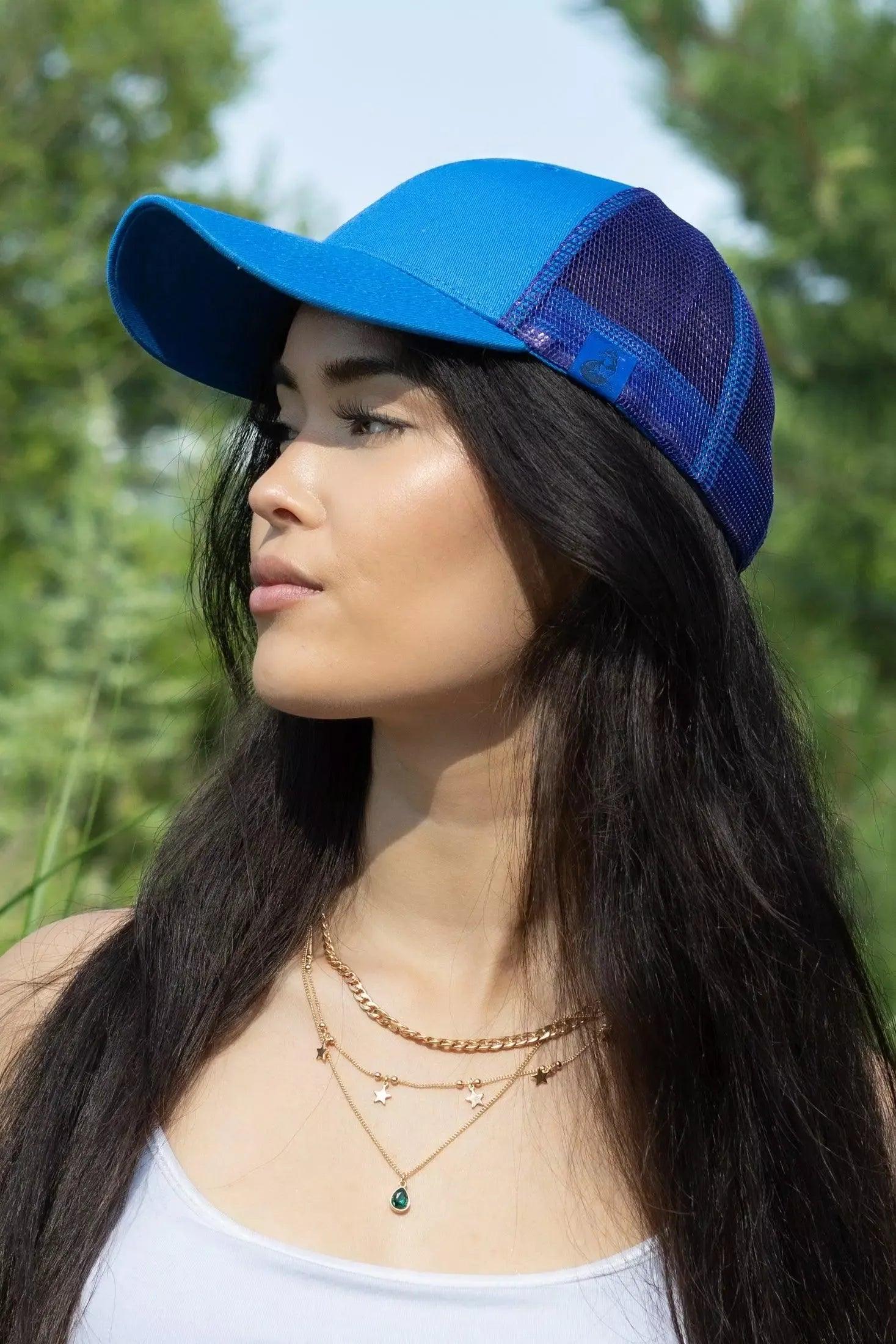 Woman wearing a blue cap with a mesh back, standing outdoors with greenery in the background.