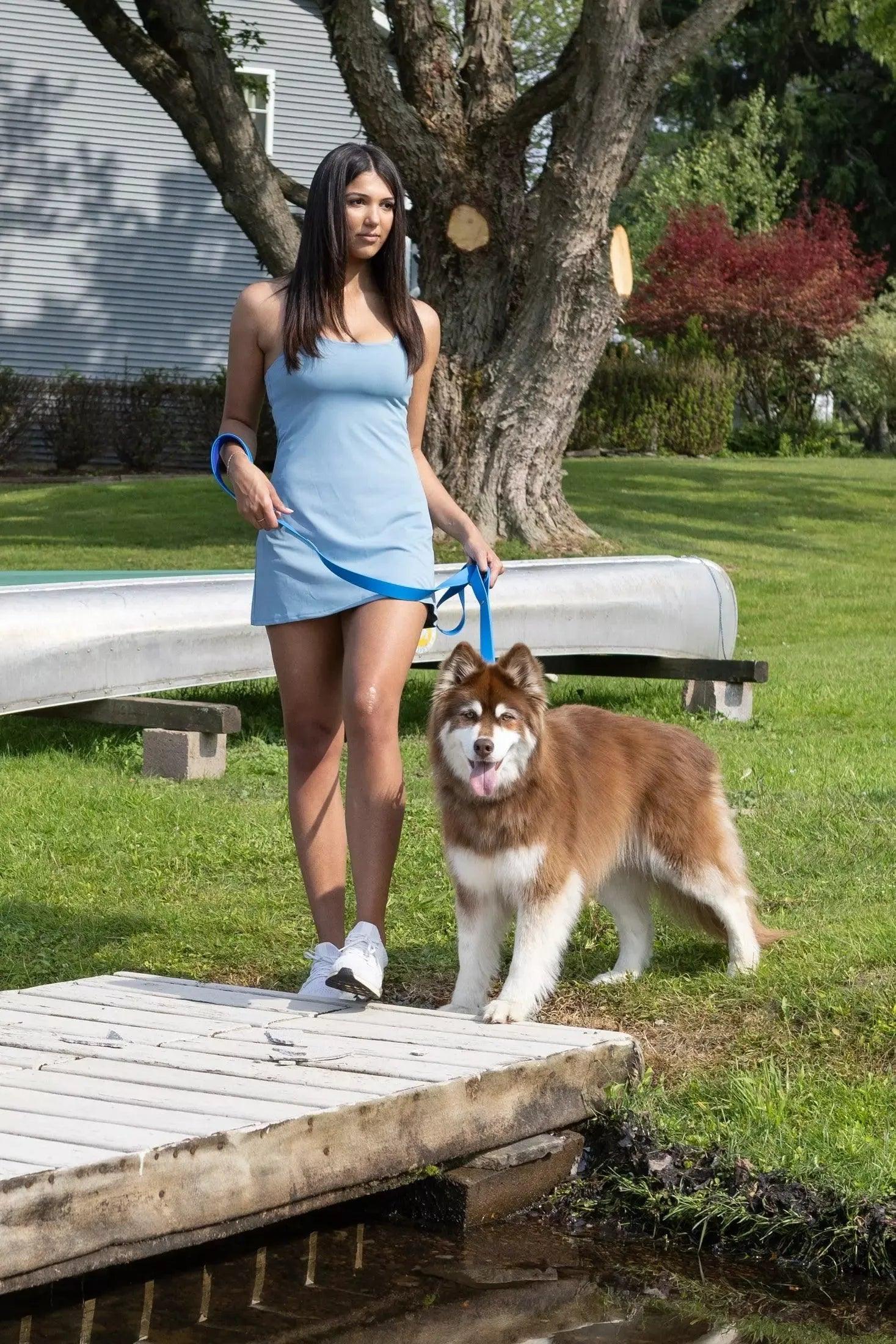 Woman in a blue dress standing with a dog on a leash in a park-like setting.