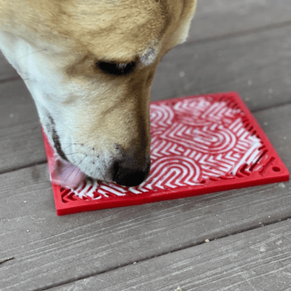 Dog licking a red lick  mat with white patterns on a wooden surface