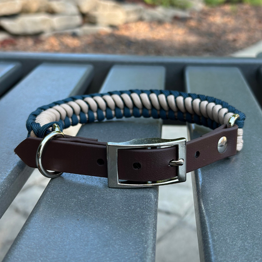 Dog collar with navy blue and light tan braided design with a dark brown BioThane strap belt buckle style silver hardware, sitting on a gray bench