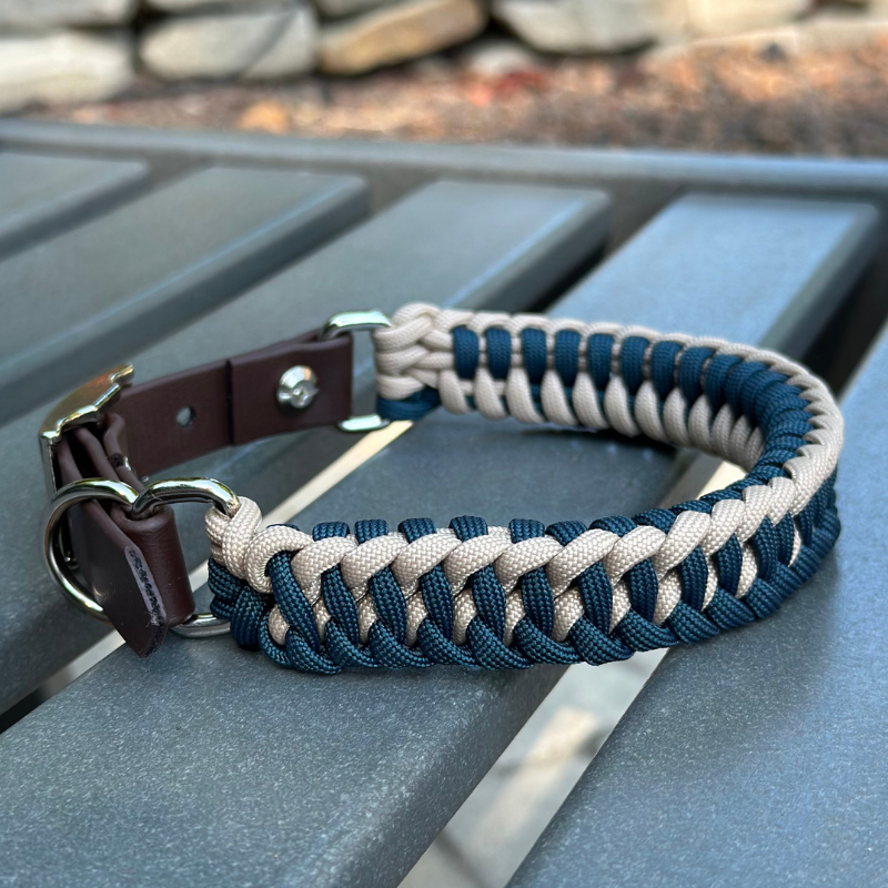 Dog collar with navy blue and light tan braided design with a dark brown BioThane strap belt buckle style silver hardware, sitting on a gray bench