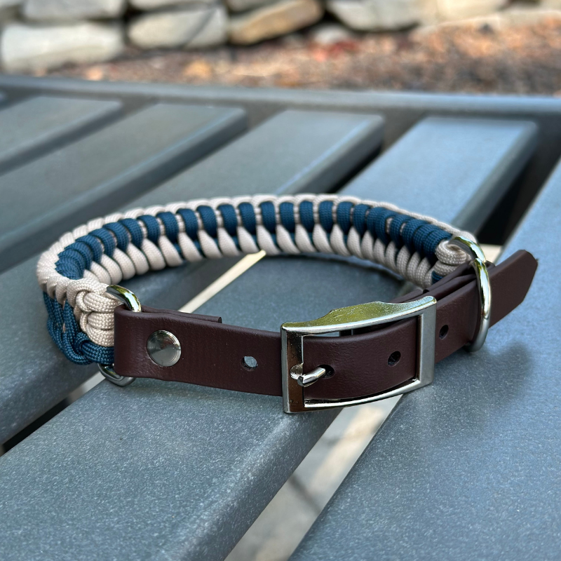 Dog collar with navy blue and light tan braided design with a dark brown BioThane strap belt buckle style silver hardware, sitting on a gray bench