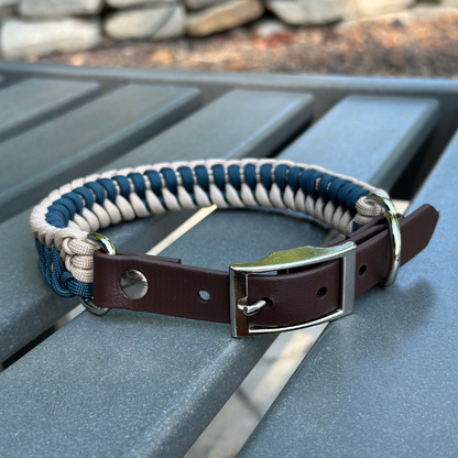 Dog collar with navy blue and light tan braided design with a dark brown BioThane strap belt buckle style silver hardware, sitting on a gray bench