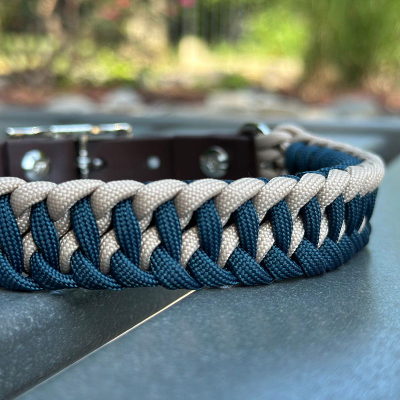 Dog collar with navy blue and light tan braided design with a dark brown BioThane strap belt buckle style silver hardware, sitting on a gray bench