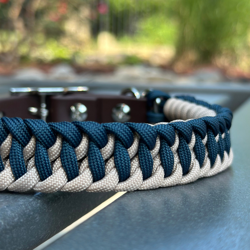Dog collar with navy blue and light tan braided design with a dark brown BioThane strap belt buckle style silver hardware, sitting on a gray bench