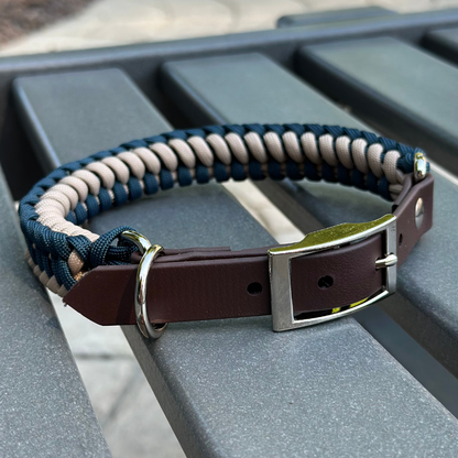 Dog collar with navy blue and light tan braided design with a dark brown BioThane strap belt buckle style silver hardware, sitting on a gray bench