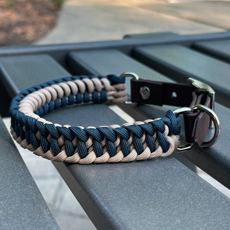 Dog collar with navy blue and light tan braided design with a dark brown BioThane strap belt buckle style silver hardware, sitting on a gray bench