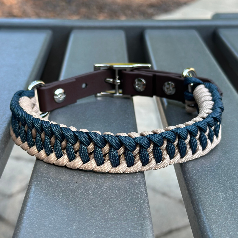 Dog collar with navy blue and light tan braided design with a dark brown BioThane strap belt buckle style silver hardware, sitting on a gray bench