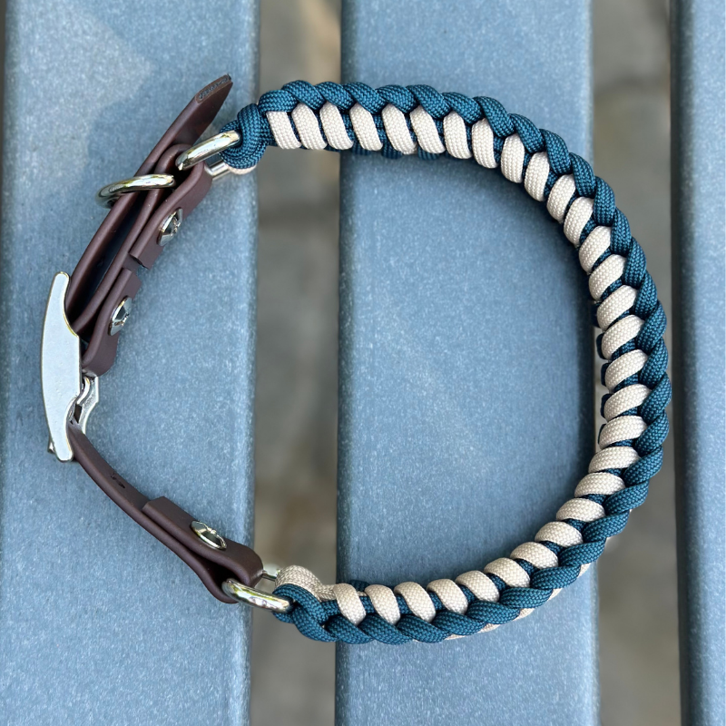 Dog collar with navy blue and light tan braided design with a dark brown BioThane strap belt buckle style silver hardware, sitting on a gray bench