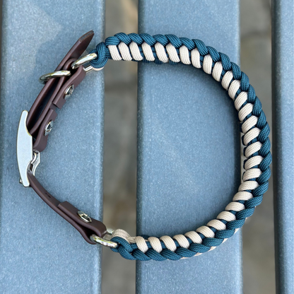 Dog collar with navy blue and light tan braided design with a dark brown BioThane strap belt buckle style silver hardware, sitting on a gray bench