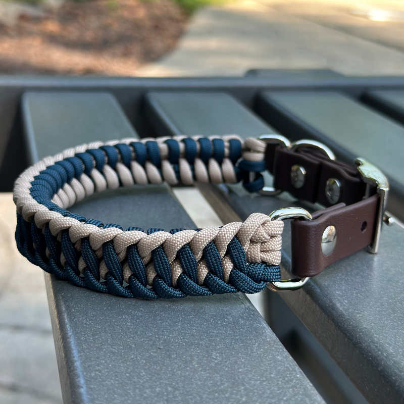 Dog collar with navy blue and light tan braided design with a dark brown BioThane strap belt buckle style silver hardware, sitting on a gray bench