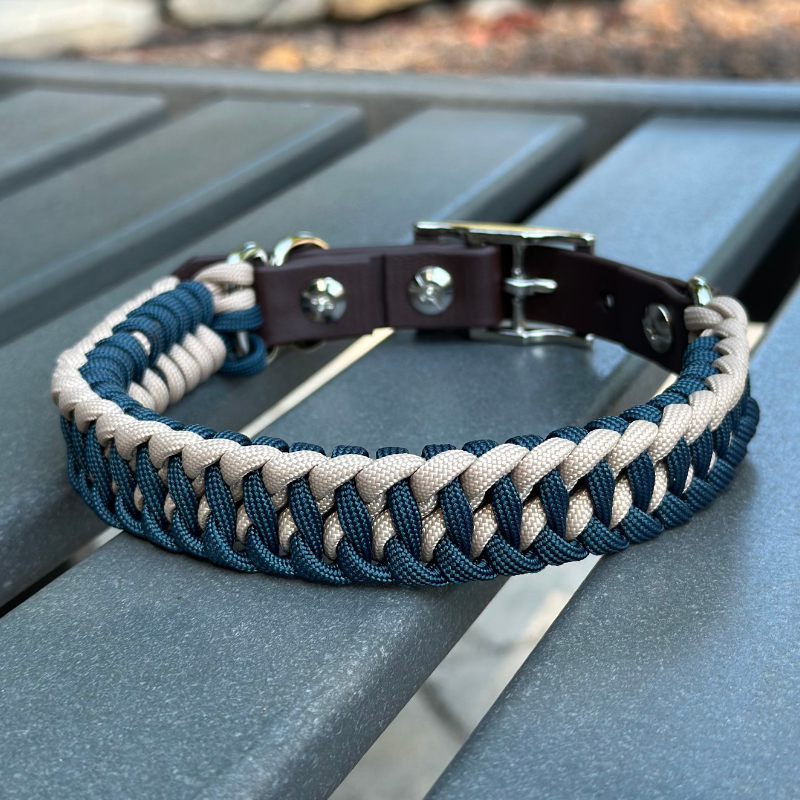 Dog collar with navy blue and light tan braided design with a dark brown BioThane strap belt buckle style silver hardware, sitting on a gray bench