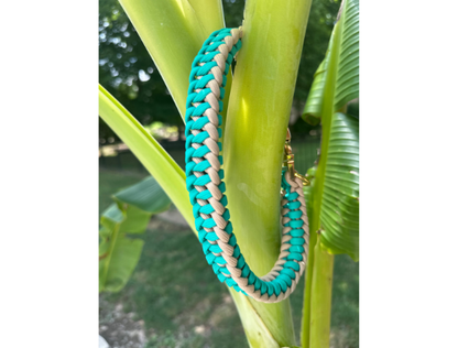 Turquoise and beige braided dog collar hanging on a banana leaf with a blurred green background
